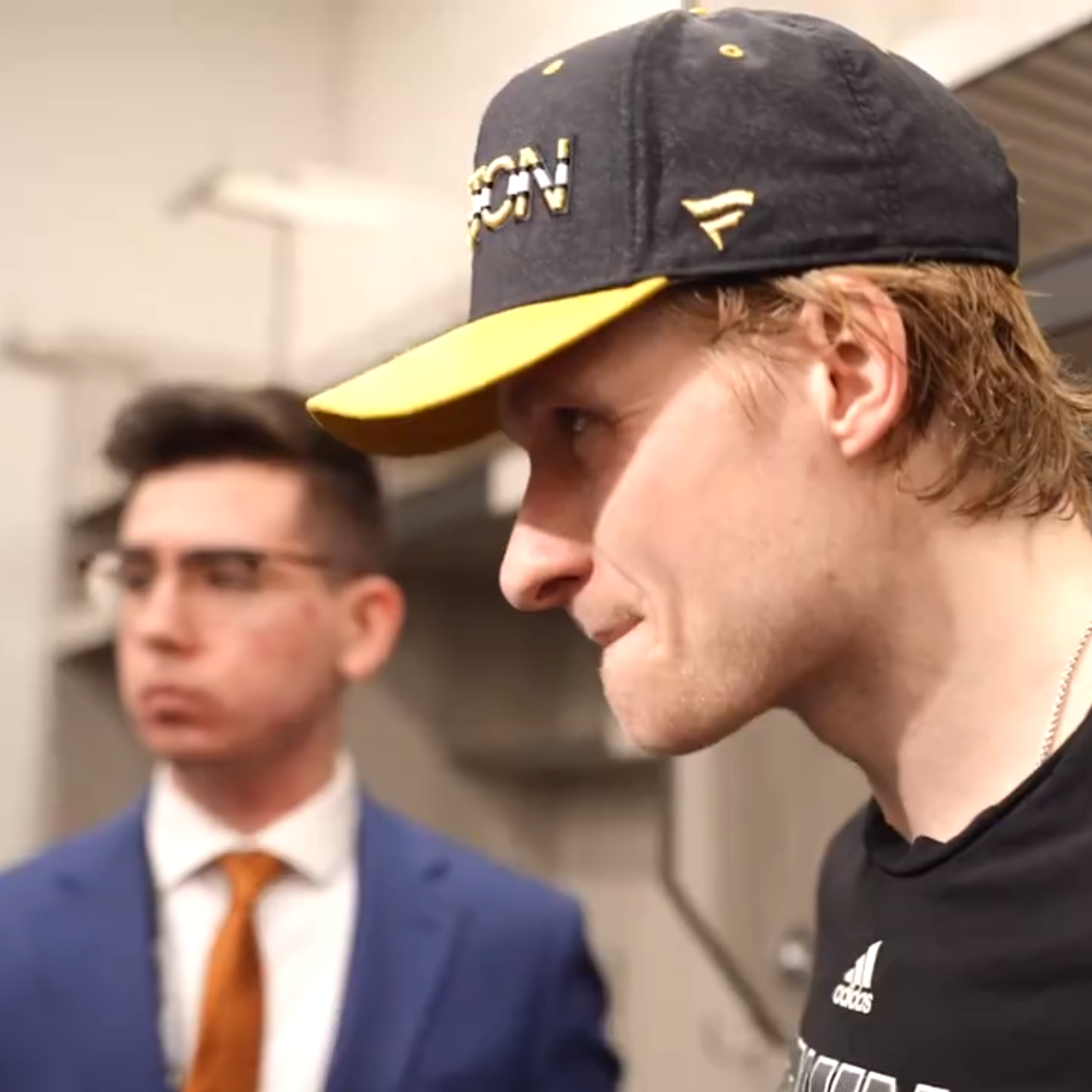Cody, a white male in a white shirt, blue blazer, and orange tie, stands behind Boston Bruins player Danton Heinen, a white male wearing a black Bruins hat, during a postgame media scrum in the visiting locker room at Wells Fargo Center in Philadelphia, Pennsylvania.