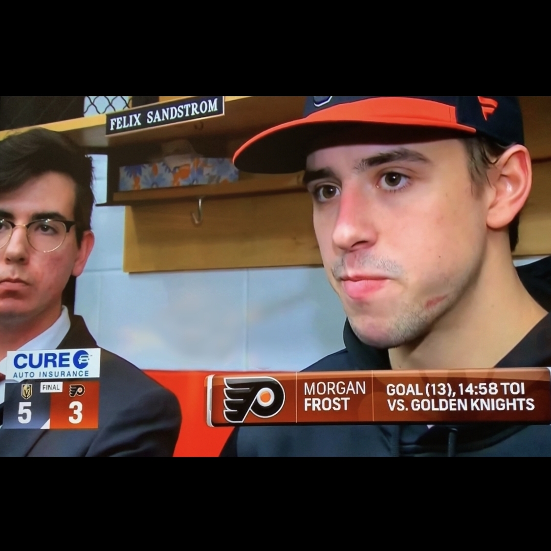 Cody, a white male with brown hair and glasses, wearing a white shirt, dark tie, and dark colored blazer, stands with Philadelphia Flyers forward Morgan Frost, a white male wearing a black Flyers sweatshirt and an orange-brimmed Flyers baseball cap, during a postgame media scrum in the Philadelphia locker room at Wells Fargo Center in Philadelphia, Pennsylvania. At the bottom of the frame, an animated on-screen chyron states that Morgan Frost scored his thirteenth goal of the season during the game and played fourteen minutes, fifty eight seconds of ice time in the Flyers' five-to-three loss against the Vegas Golden Knights.