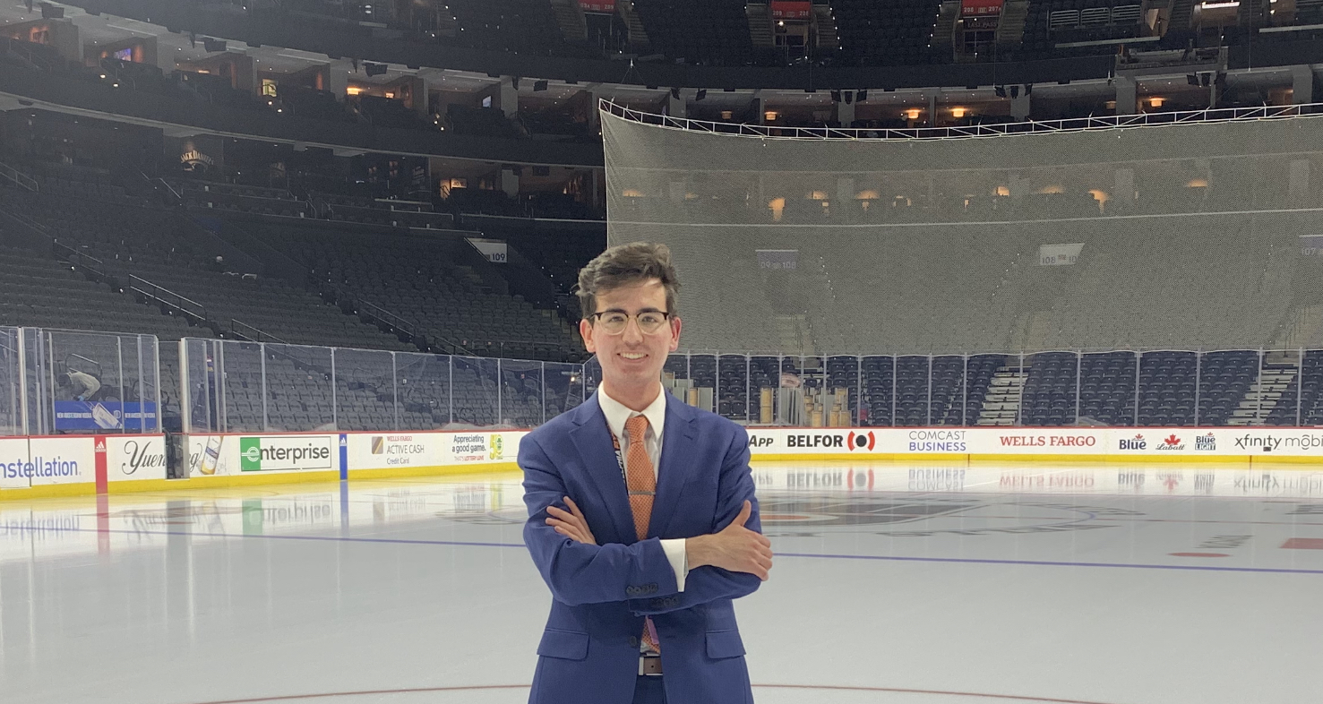 Cody, a white male with dark hair, wearing glasses, a blue blazer and orange tie, standing on the ice at the Wells Fargo Center in Philadelphia, Pennsylvania.