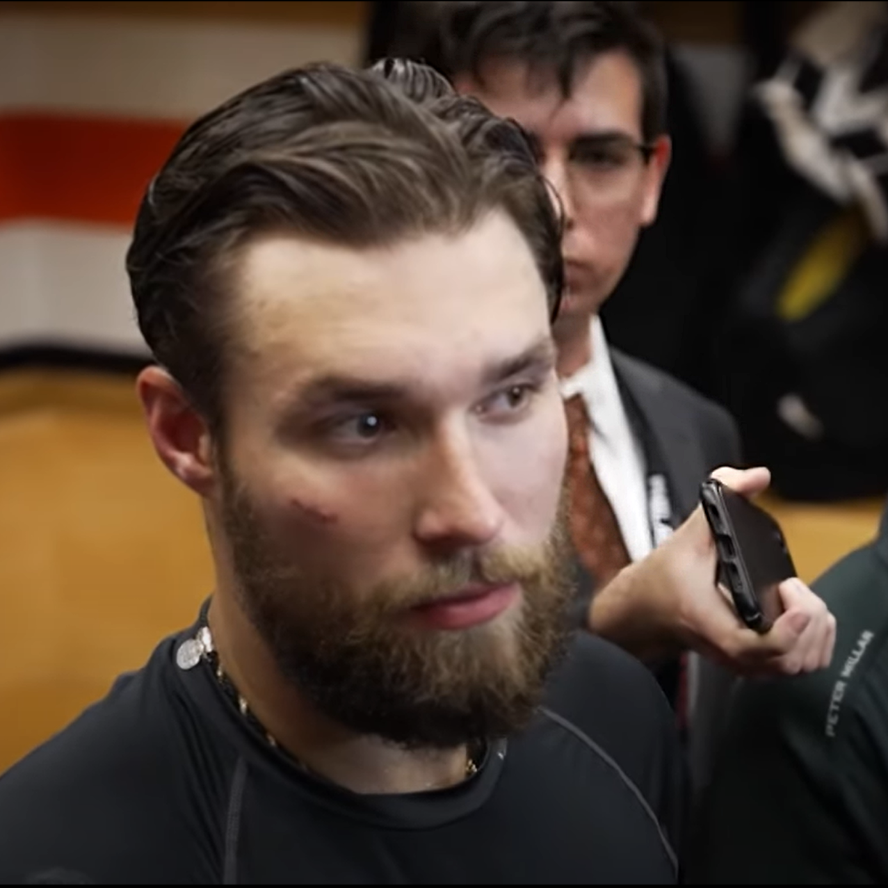 Cody, a white male with brown hair and glasses, wearing a white shirt, dark orange tie, and dark colored blazer, stands behind Philadelphia Flyers defenseman Ivan Provorov during a postgame media scrum in the Philadelphia locker room at Wells Fargo Center in Philadelphia, Pennsylvania.