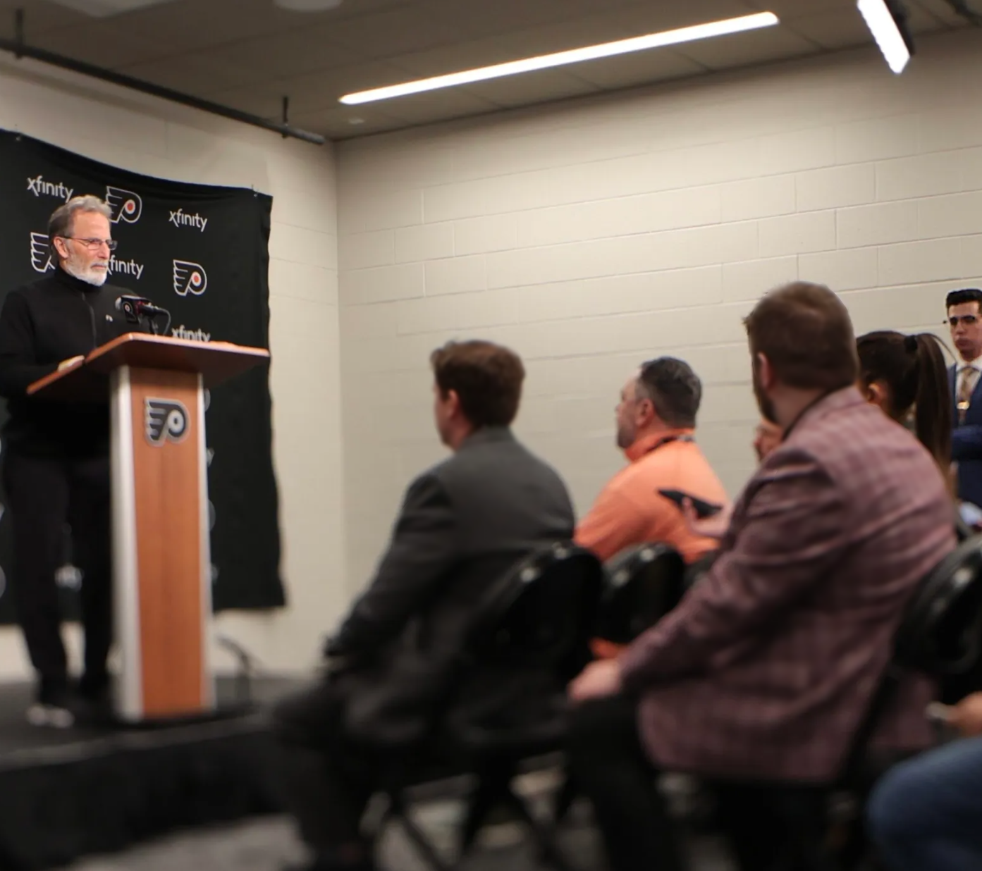 Cody, a white male in a white shirt and dark jacket, stands off to the right side of Head Coach John Tortorella's postgame media availability in the Zack Hill Media Room located at Wells Fargo Center in Philadelphia, Pennsylvania.