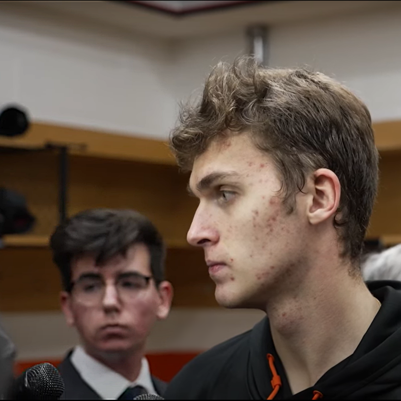 Cody, a white male with brown hair and glasses, wearing a white shirt, dark tie, and dark colored blazer, stands behind Philadelphia Flyers defenseman Egor Zaumula during a postgame media scrum in the Philadelphia locker room at Wells Fargo Center in Philadelphia, Pennsylvania.