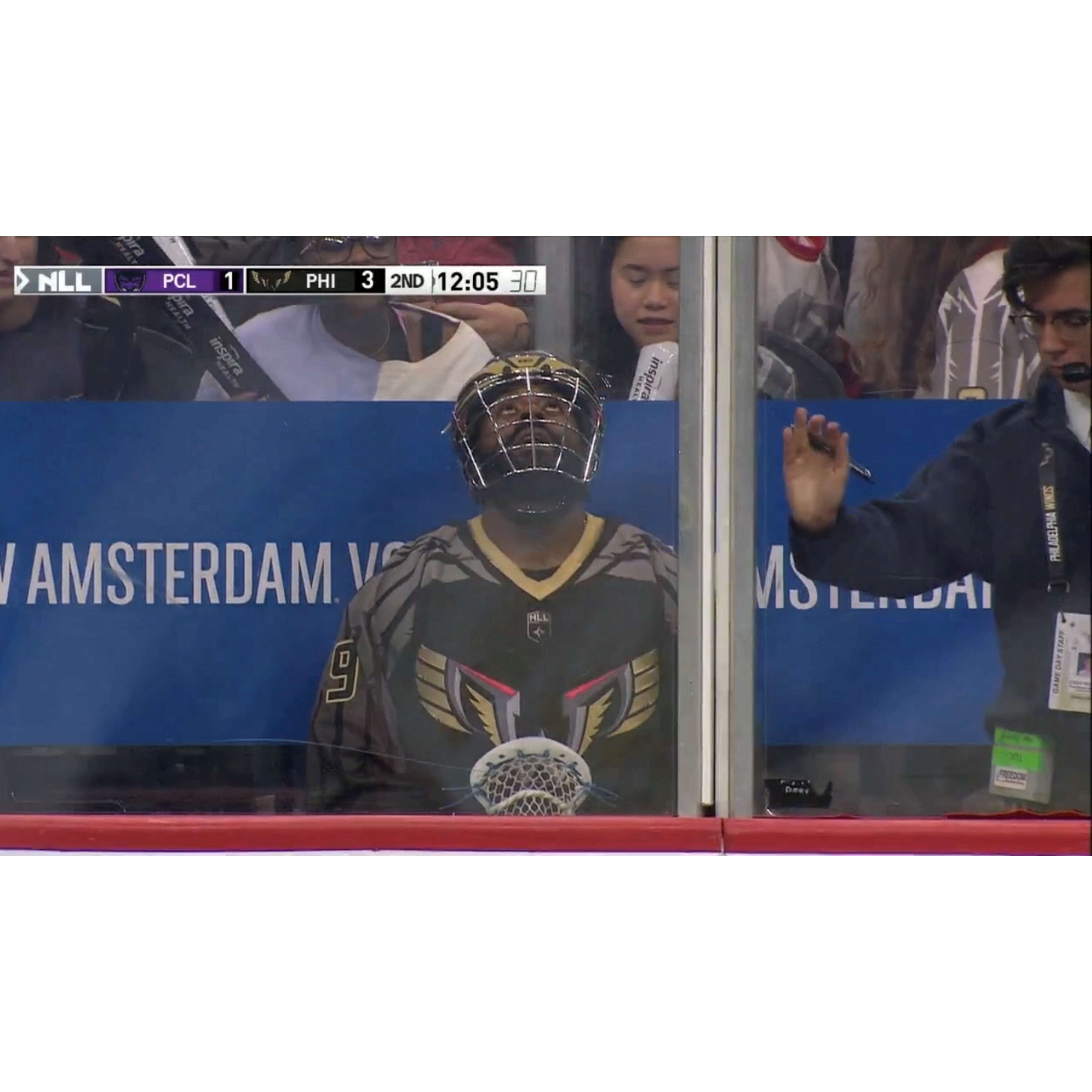Cody, a white male with brown hair, glasses, wearing a dark blue quarter zip jacekt, is seen from across the field as he closes the penalty box door during a live Philadelphia Wings versus Panther City Lacrosse club game at Wells Fargo Center. Philadelphia Wings player Trevor Baptiste, wearing a black and gold lacrosse helmet with a black Wings Lacrosse jersey, is seated behind Cody in the penalty box.